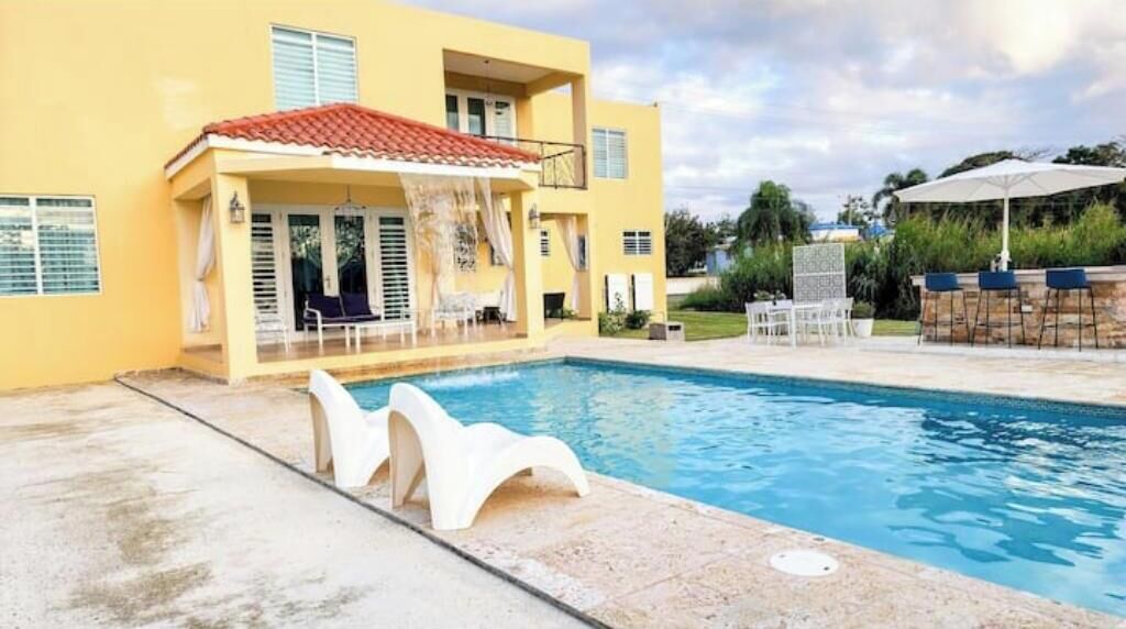 A yellow building with a swimming pool, outdoor seating, and a covered patio area.