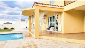A yellow building with a covered patio, a pool, and a chair.