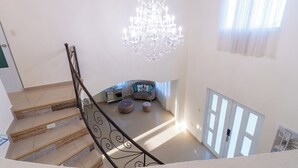Un escalier orné d’une rampe en fer forgé mène à un salon agrémenté d’un lustre, d’un ensemble de canapés et d’une fenêtre avec des rideaux légers.