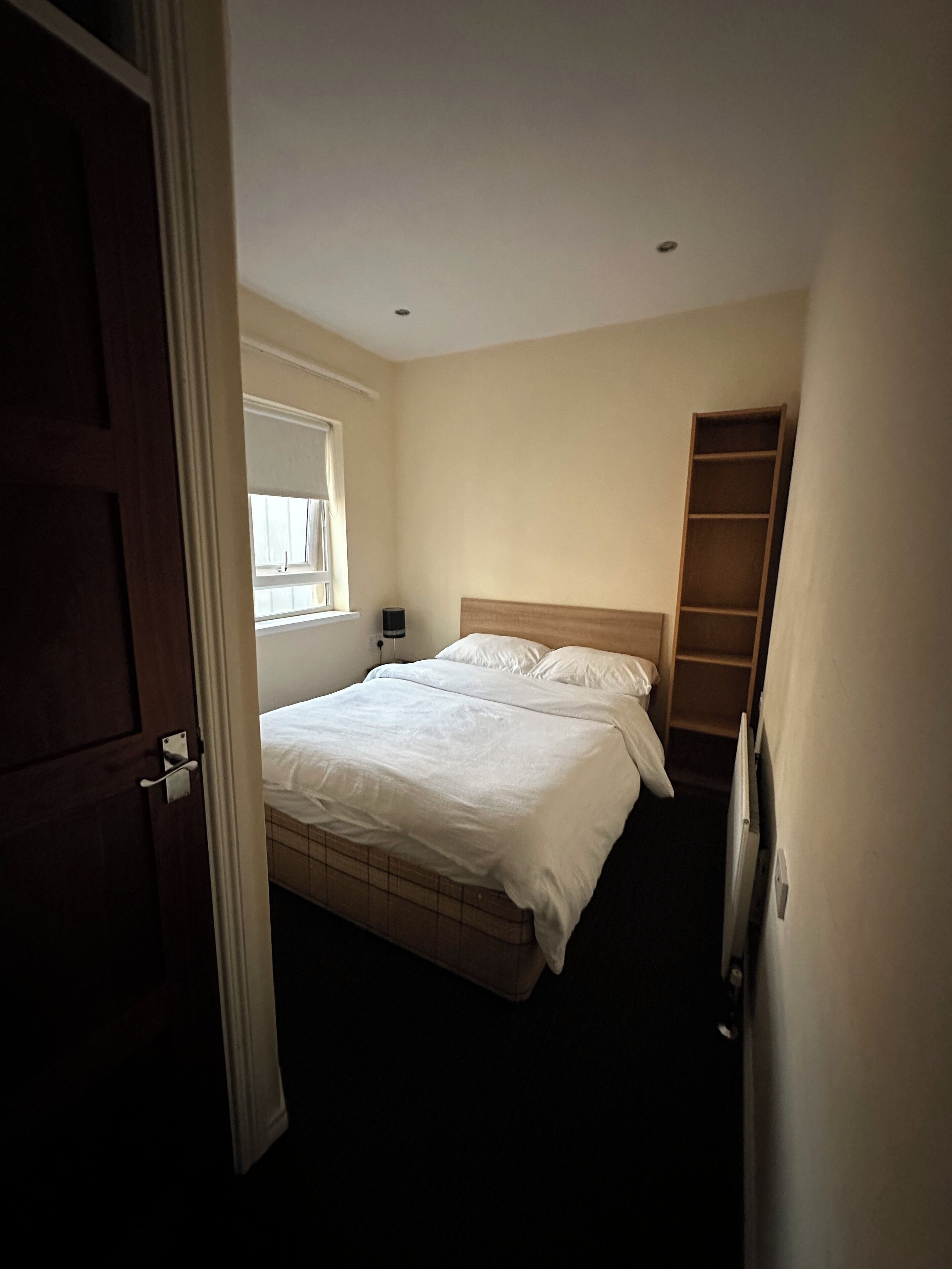 Un dormitorio con cama, ventana, mesita de noche y estante de madera.