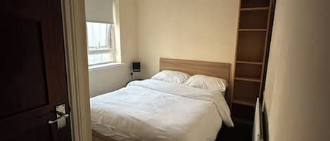Un dormitorio con cama, ventana, mesita de noche y estante de madera.