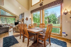 Un espace repas avec une table et des chaises en bois, de grandes fenêtres donnant sur un paysage verdoyant, et un salon confortable en arrière-plan.