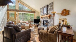 A spacious living room with a stone fireplace, leather sofas, and a large window offering a view of the outdoors. - Elk River Cabin – On Big Thompson River, Near RMNP, Estes Park – 23-ZONE3554 (Estes Park)