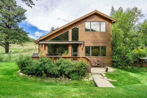 Une cabane en rondins à deux étages, dotée d’une terrasse, entourée d’une végétation luxuriante et sous un ciel dégagé.