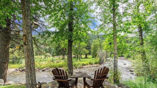 A rustic outdoor seating area with two wooden chairs and a table, surrounded by trees and a stone pathway. - Elk River Cabin – On Big Thompson River, Near RMNP, Estes Park – 23-ZONE3554 (Estes Park)