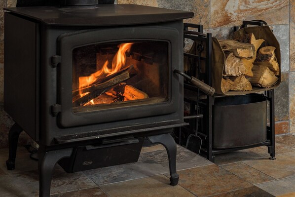 Ein schwarzer Kaminofen mit sichtbaren Flammen, ein Metallholzhakenzähler und eine Steinsäule im Hintergrund.