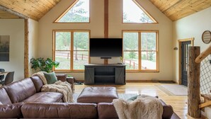 Een ruime woonkamer met een grote televisie, houten plafond en grote ramen.