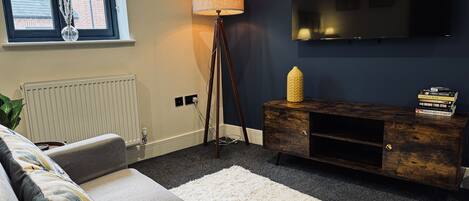 Ein modernes Wohnzimmer mit grauem Sofa, einem weißen Teppich, einer Stehlampe, einem an der Wand befestigten Fernseher und einer Holzkommode mit Büchern.