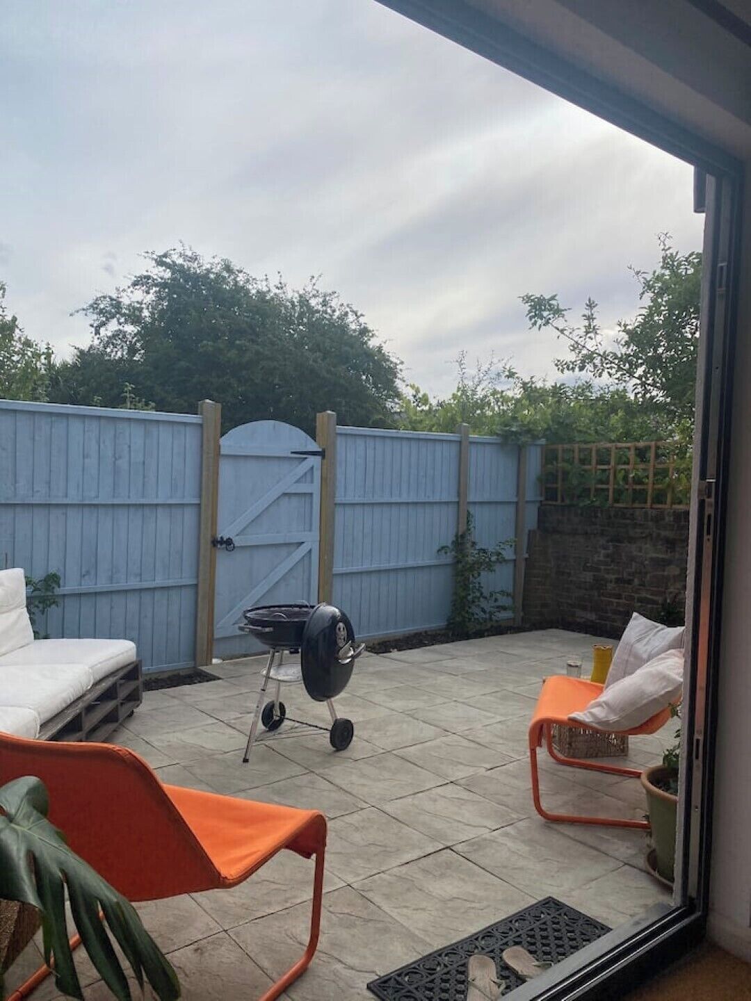 Eine Terrasse mit Grill, zwei orangefarbenen Stühlen, einem weißen Sofa und einem blauen Zaun.