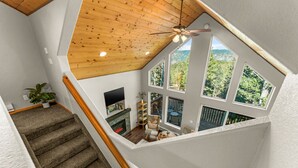 A spacious loft with a high ceiling, a fireplace, a TV, and a balcony with a view of the surrounding forest. - Epic Views, Modern Cabin, Hot tub, Family & Pets (Woodland Park)