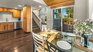 A modern kitchen with wooden cabinets, a dishwasher, and a dining area with a table set for two. - Epic Views, Modern Cabin, Hot tub, Family & Pets (Woodland Park)