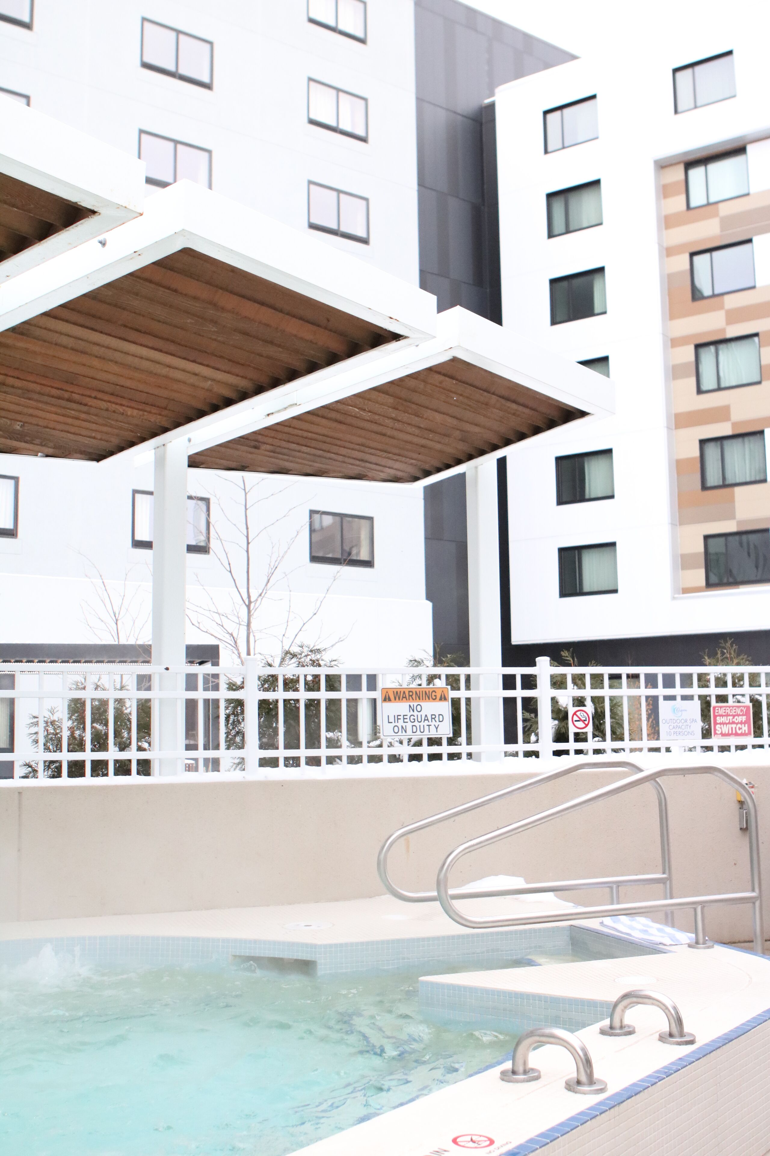 A swimming pool with a wooden deck overhead, surrounded by a white fence with a lifeguard sign, and a modern building in the background.
