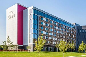 Un moderno edificio de hotel con una fachada llamativa en rosa y blanco, varios pisos y un césped bien cuidado con árboles jóvenes.