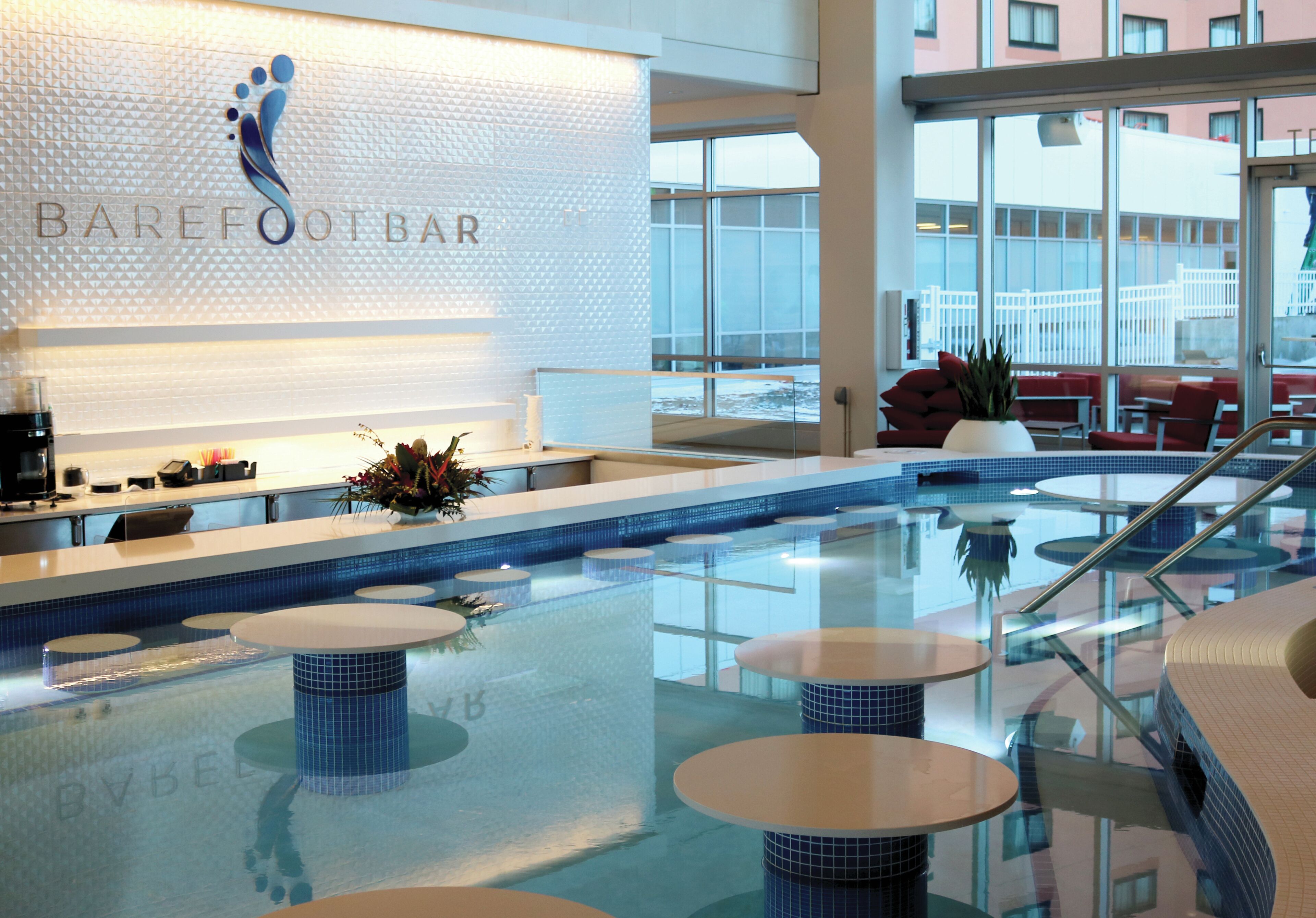 A modern indoor pool area with a Barefoot Bar sign, multiple seating areas, and a view of the outside.