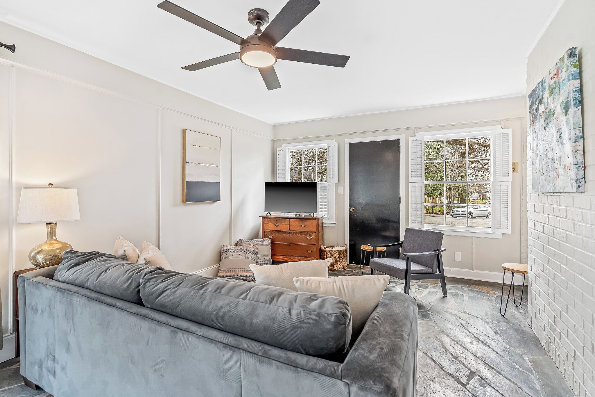A living room with a grey sofa, a ceiling fan, a TV on a wooden cabinet, and a window with shutters.