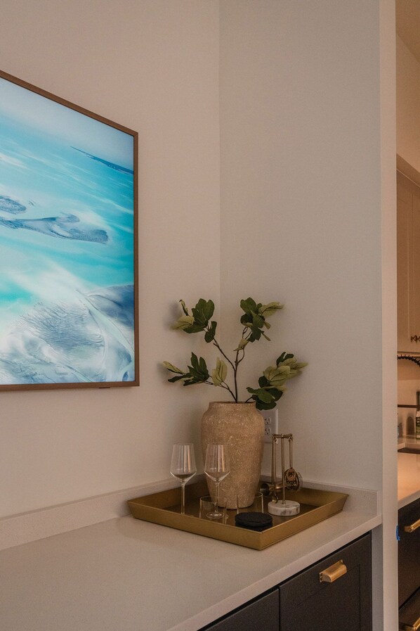 A modern kitchen with a large painting, a vase with green leaves, and two wine glasses on a tray.