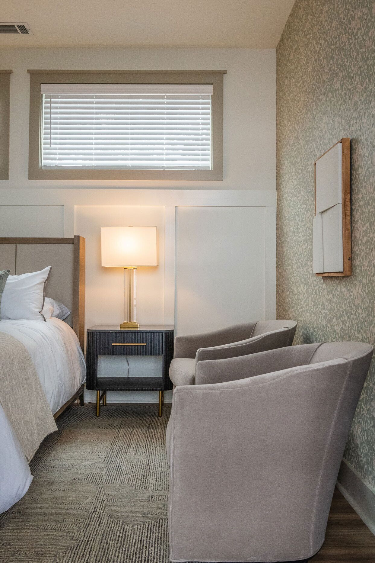 Een hotelkamer met een bed, nachtkastje met lamp, stoel en een klein tafeltje.