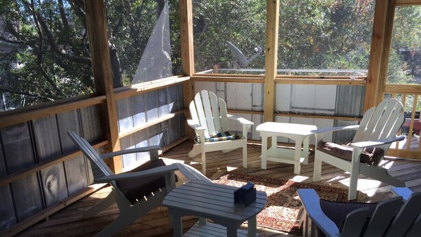 Eine überdachte Terrasse im Freien mit Adirondack-Stühlen und einem kleinen Tisch, umgeben von Holzbalkonen und Blick auf Bäume.