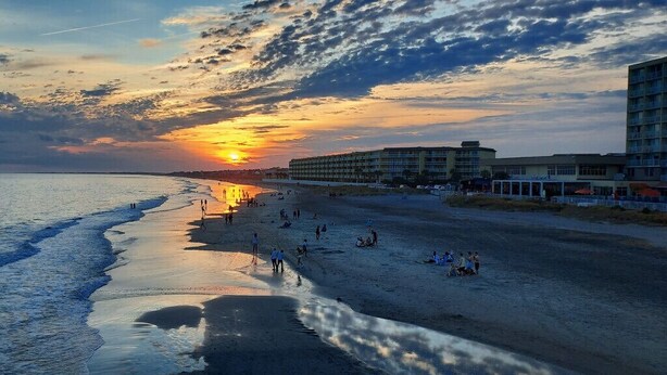 A beach with a sunset, people on the sand, and buildings in the background. - “Crescent Moon” - 5BR – established, top rated, dog friendly, 5 min walk to beach! (Folly Beach)