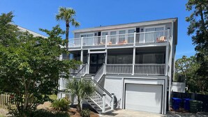 A two-story residential building with a front garage, a balcony on the upper floor, and a small front yard with a palm tree and shrubs. - “Crescent Moon” - 5BR – established, top rated, dog friendly, 5 min walk to beach! (Folly Beach)