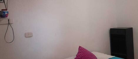 A small, simple room with a bed, a chair, a TV, and a wall-mounted air conditioner.