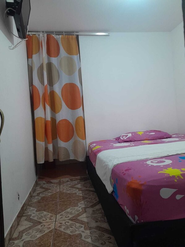 Una habitación pequeña con cama individual, cortina estampada, ropa de cama colorida y baldosas de suelo con diseño.