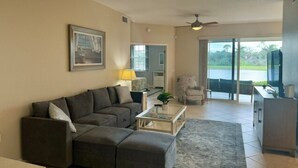A living room with a grey sectional sofa, a glass coffee table, and a view of a balcony with a pool. - Beautiful Relaxing condo with a Lake/Golf Course view 8 miles from beach! (Fort Myers)
