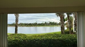 A view from a room with a large window overlooking a lake, surrounded by greenery and palm trees. - Beautiful Relaxing condo with a Lake/Golf Course view 8 miles from beach! (Fort Myers)