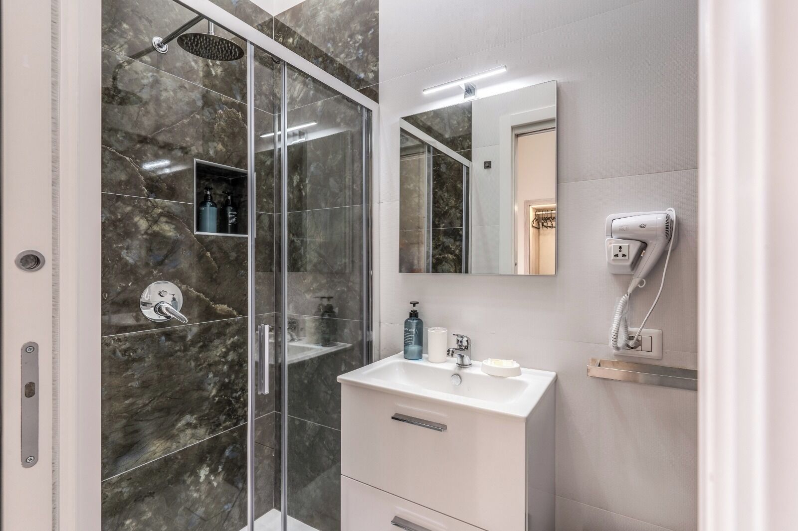 A modern bathroom with a glass-enclosed shower, a white vanity with a mirror, and a hairdryer on the wall.