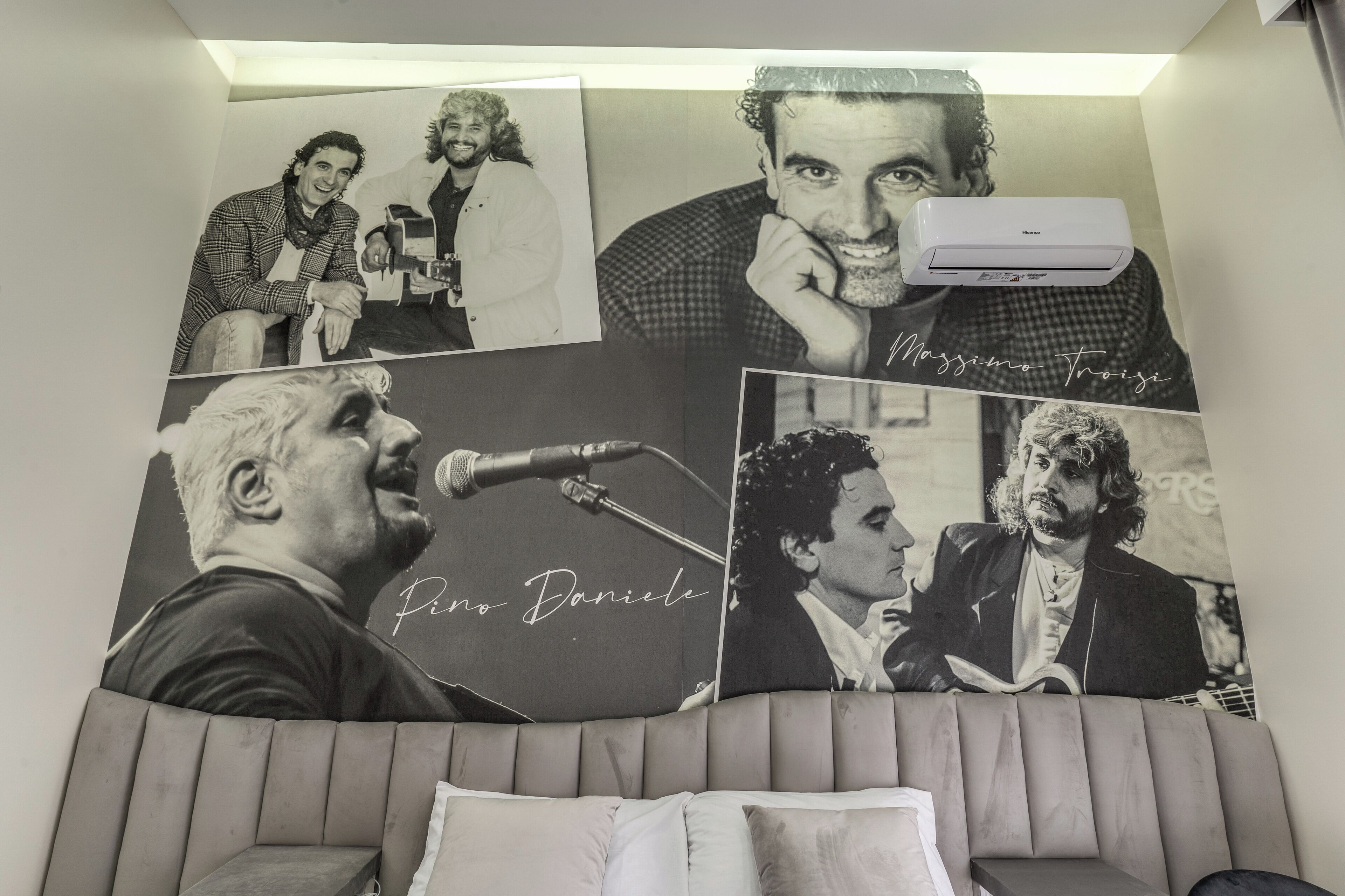 A room with a large wall of framed black and white photographs of musicians, a white air conditioner, and a tufted headboard.