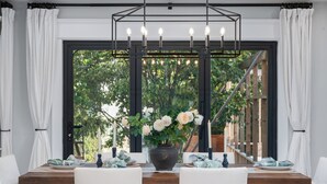 A dining area with a table set for two, a vase with flowers, and a view of greenery through a large window. - Family getaway.Commuter's Dream.Weddings&Events.Content House.Luxury Smart Home (Larchmont)