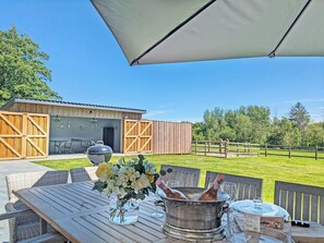 Een terras met een tafel, een metalen emmer met flessen, en een gebouw met houten deuren en een hek op de achtergrond.