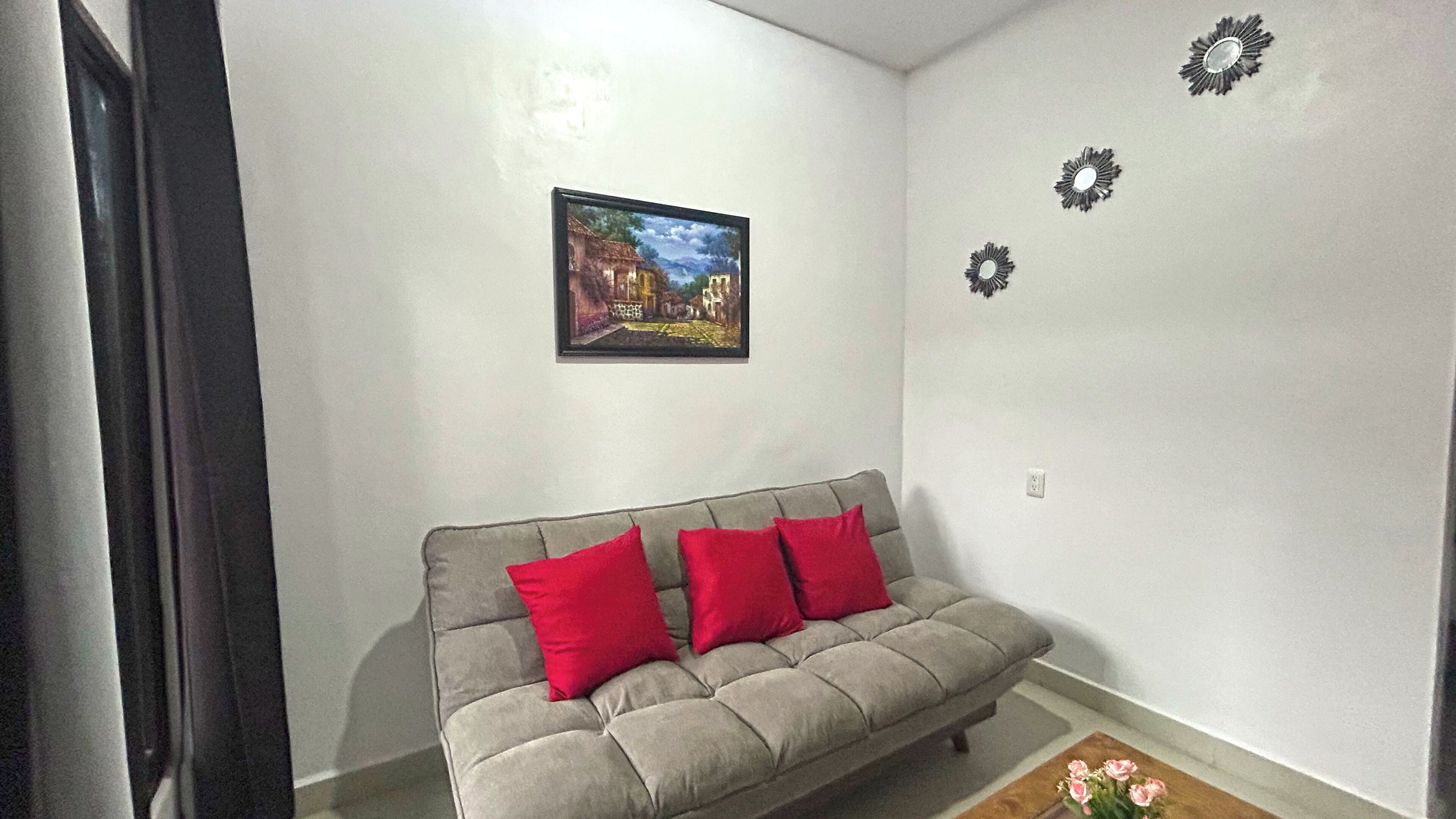 Un salón con sofá gris, cojines rojos, un cuadro enmarcado en la pared y espejos decorativos.