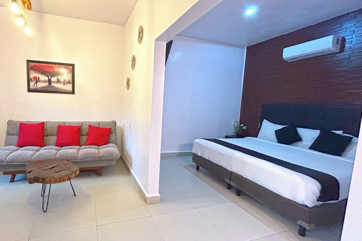 A modern bedroom with a bed, a sofa with red cushions, a small wooden stool, and a painting on the wall.