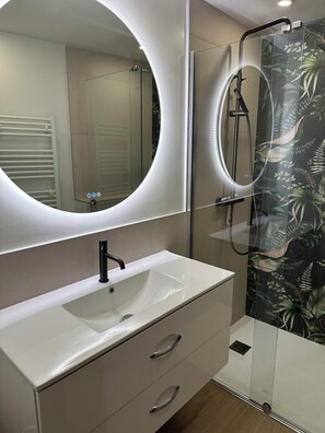 Un baño con un tocador blanco, un espejo redondo y una ducha con puerta de cristal.
