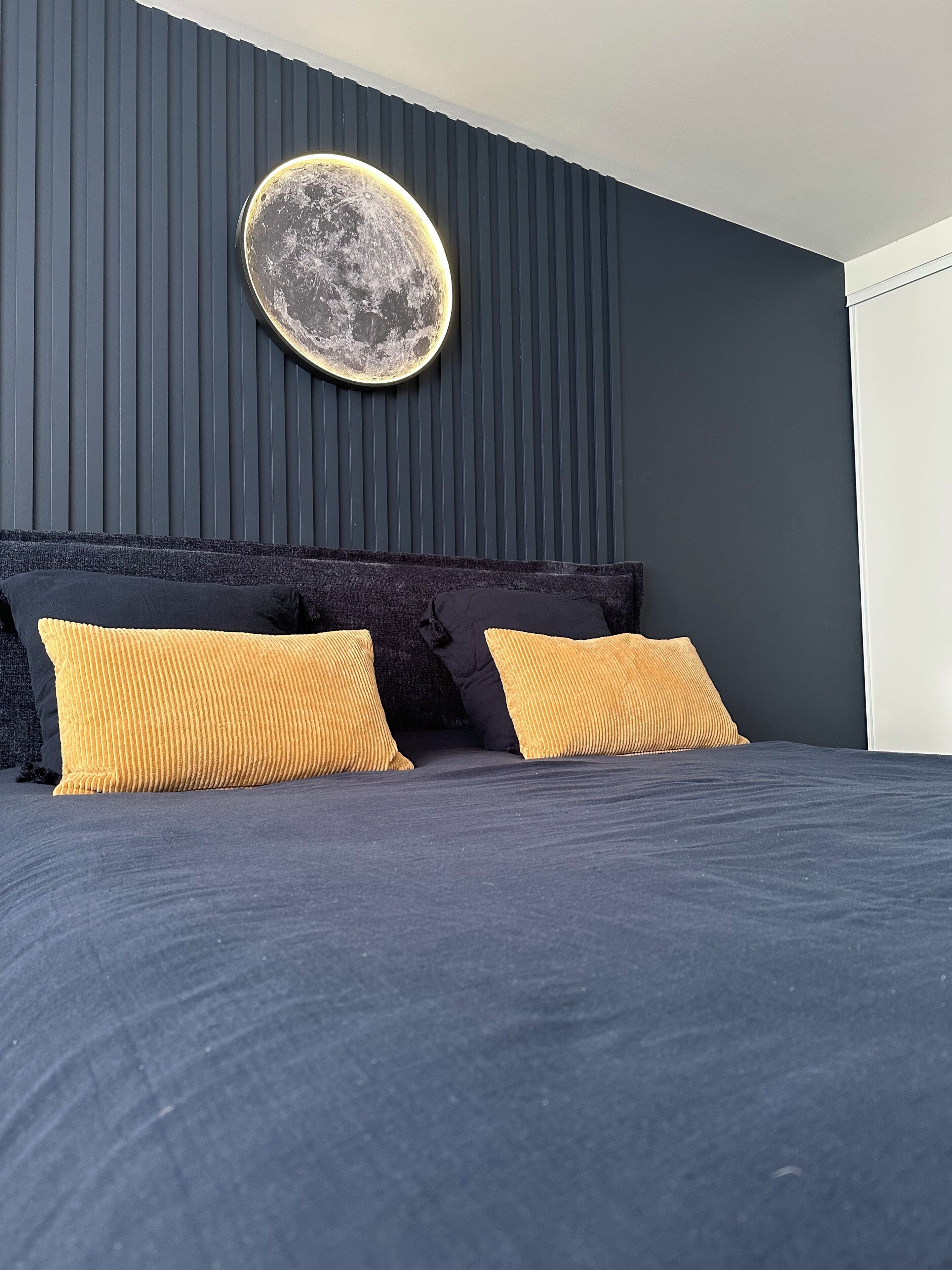 Una cama con ropa oscura, dos almohadas amarillas y una decoración circular de luna montada en la pared.