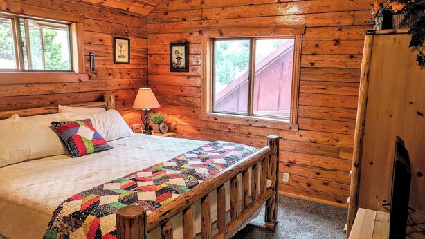 Ein Schlafzimmer einer Holzhütte mit Bett, Nachttisch, Lampe, Fernseher auf einem Schrank und einem Fenster mit Blick auf Bäume.