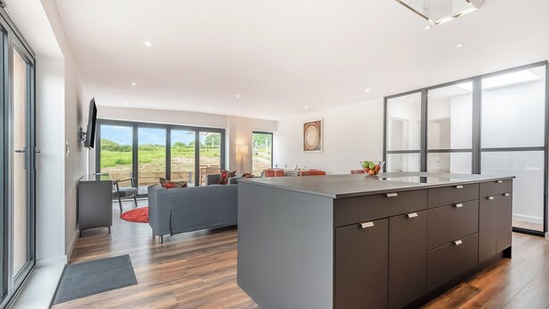 A modern kitchen with a large island, a dining area with a red rug, and large windows offering a view of the outdoors.