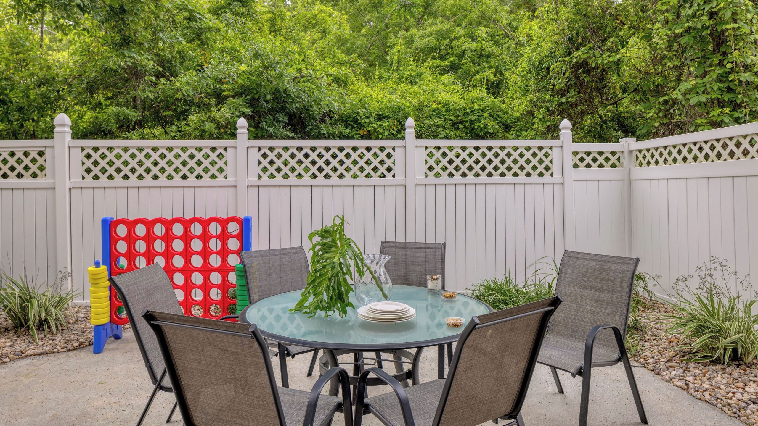 An outdoor dining area with a round glass table, four chairs, and a white picket fence.