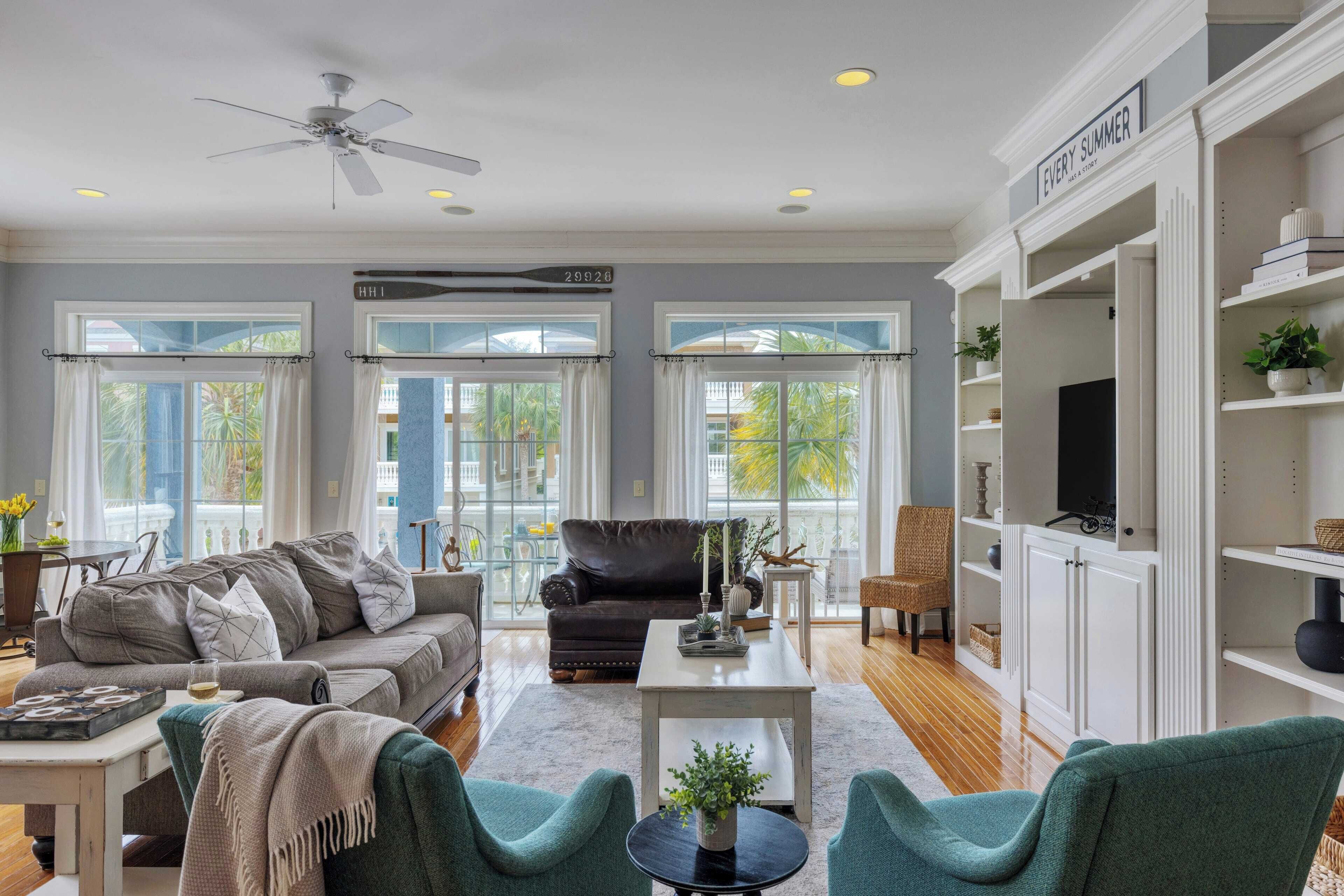 A spacious living room with a grey sofa, teal armchairs, a coffee table, and a TV on a white cabinet.