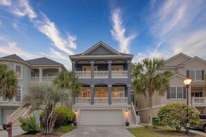 A two-story residential building with a balcony, a garage, and a well-maintained lawn with palm trees. - Crabline Getaway by Avantstay Private Pool + Hot Tub Easy Beach Access (Hilton Head Island)