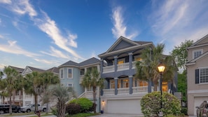 A multi-story residential building with a balcony, garage, and surrounding palm trees. - Crabline Getaway by Avantstay Private Pool + Hot Tub Easy Beach Access (Hilton Head Island)
