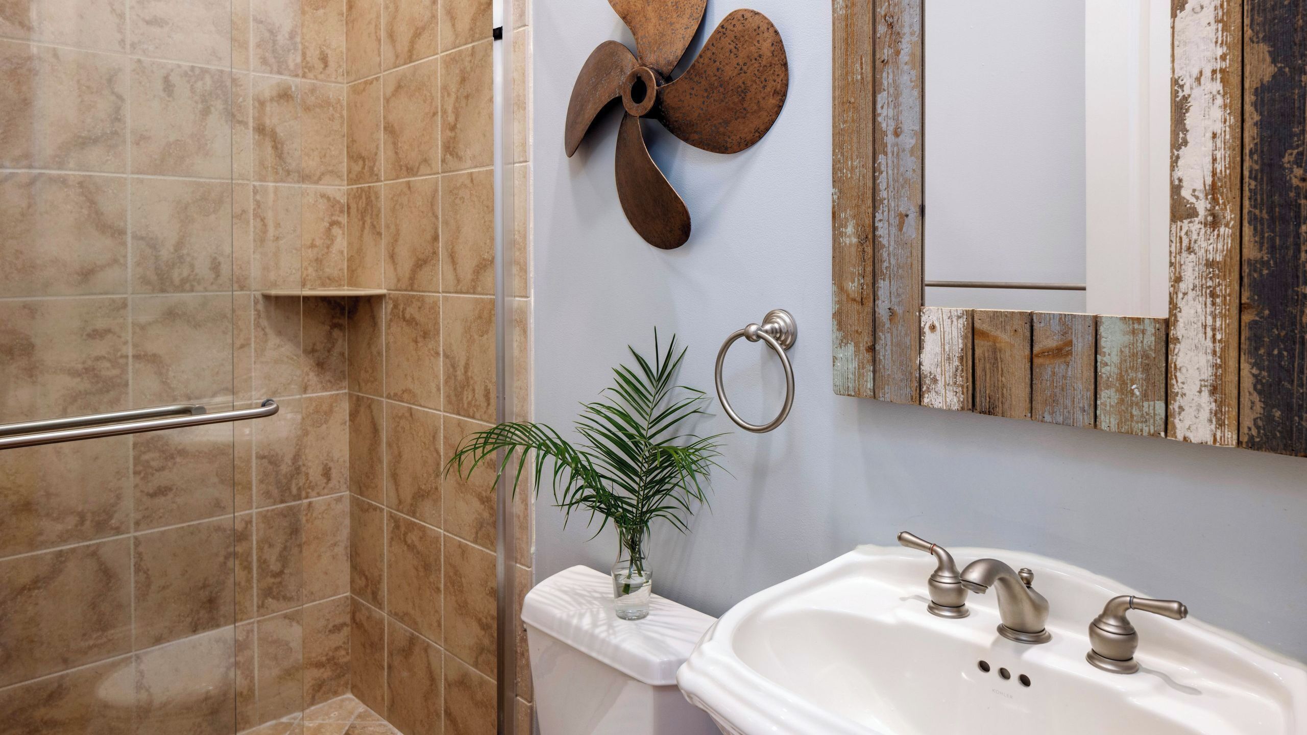 A bathroom with a shower, a sink with two faucets, a towel rack, and a decorative fan.