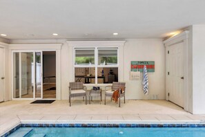 Un espace aménagé près de la piscine avec une table et des chaises, une cuisine visible à travers une porte coulissante, et un panneau indiquant où trouver les serviettes.