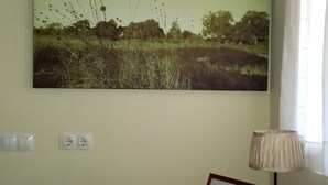 A room with a wall-mounted painting of a landscape, a lamp, and a table with a framed note. - La Canal (Santillana del Mar)