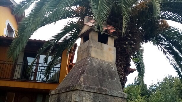 A stone structure with a chimney, surrounded by palm trees and a yellow building with a balcony. - La Canal (Santillana del Mar)