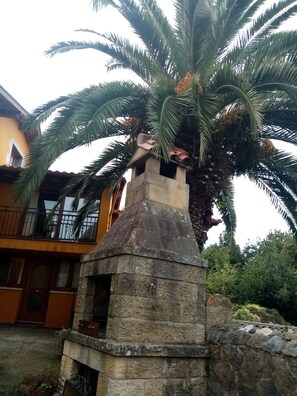 A stone structure with a chimney, surrounded by palm trees and a yellow building with a balcony. - La Canal (Santillana del Mar)