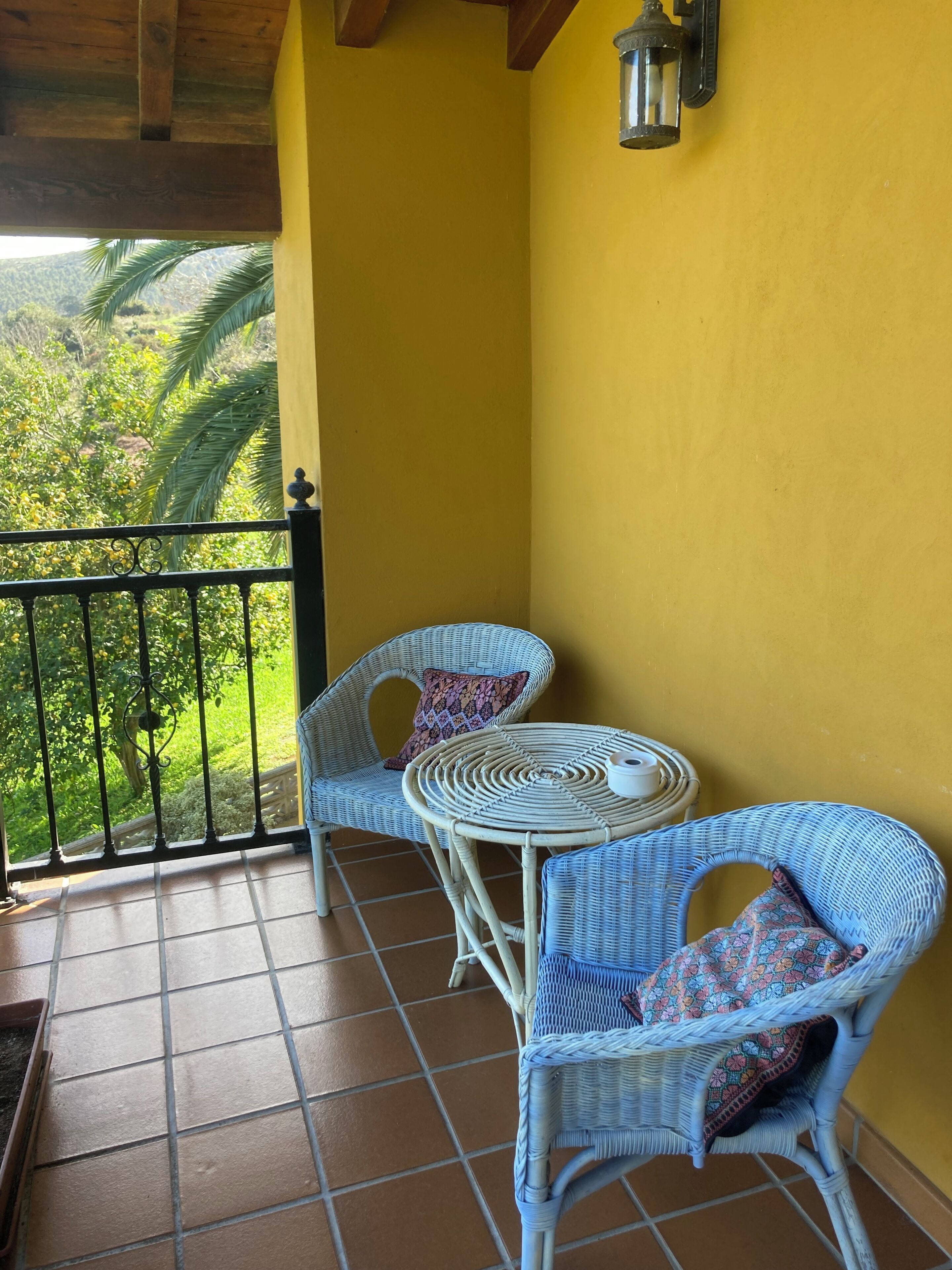 A balcony with two wicker chairs and a small round table, yellow walls, and a view of greenery.