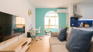 A modern living room with a flat-screen TV, a wooden TV stand, a sofa with cushions, a small table with a chair, and a kitchen area with a stove and a window.