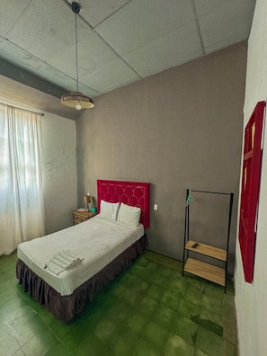 A room with a bed, a nightstand, a hanging light fixture, a window with curtains, and a red door.